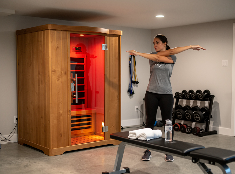 Person stretching forearms next to a home infrared sauna in a basement gym
