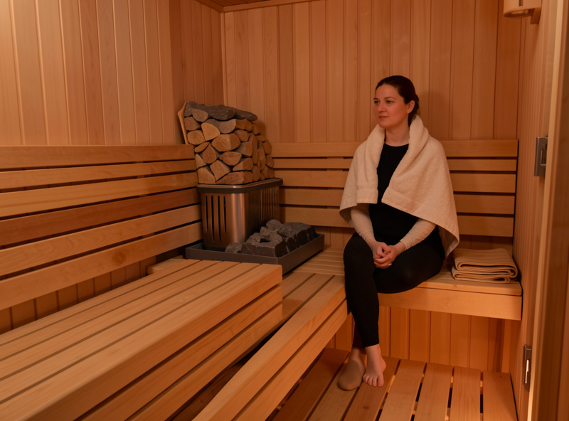 A person seated at the cooler far end of a hemlock sauna bench away from the heater