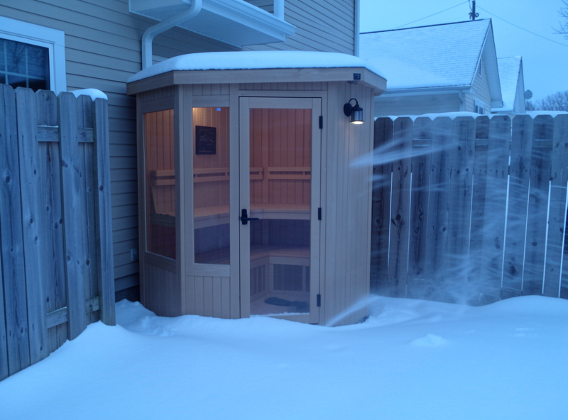Outdoor infrared sauna cabin tucked against a house wall and fence as a windbreak in blowing snow